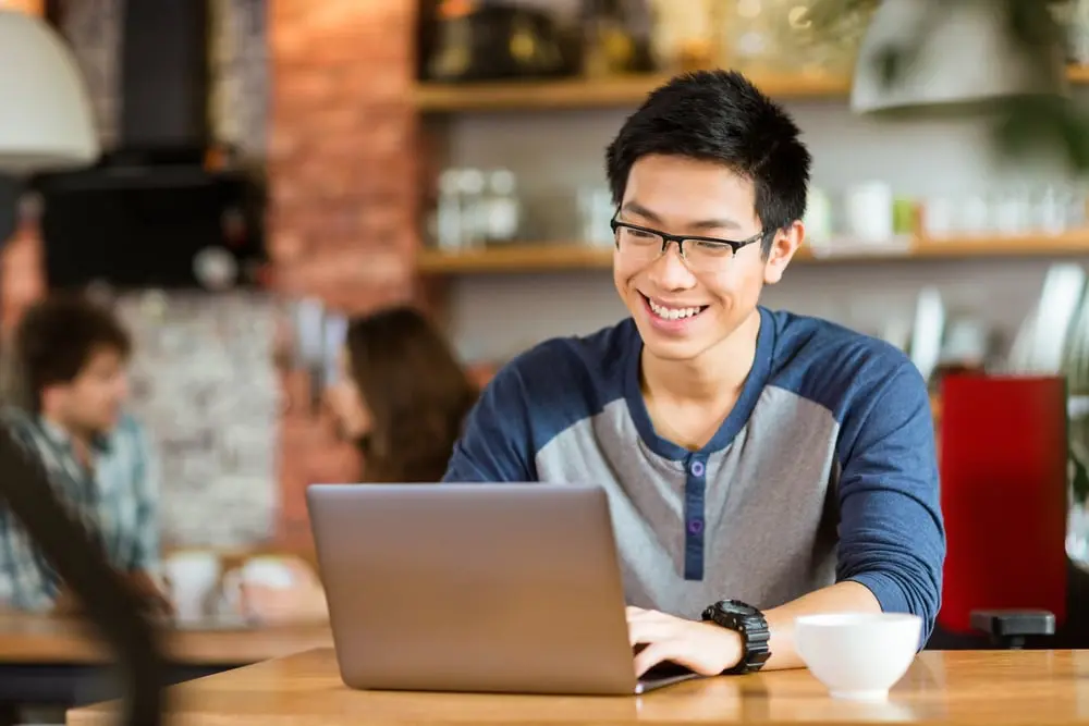 Happy cheerful young asian male in glasses smiling and using laptop in cafe_compressed